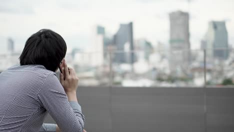 Junge-asiatische-Geschäftsmann-mit-Smartphone-für-Business-Talk-auf-Bürogebäude-Dachterrasse-mit-Blick-auf-die-Stadt-im-Hintergrund-sitzt.-Kommunikation-und-Corporation-Geschäftskonzepte.