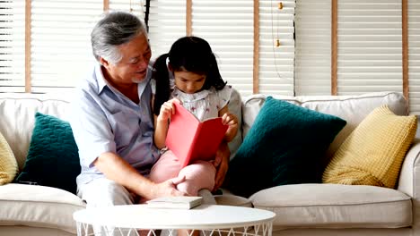 Senior-man-and-little-girl-reading-book-together-at-home.-people-with-family,-lifestyle,-education-concept.-4k-resolution.