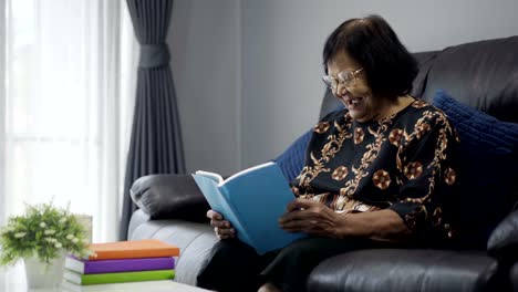 Senior-mujer-leyendo-un-libro-y-riendo-en-la-sala-de-estar