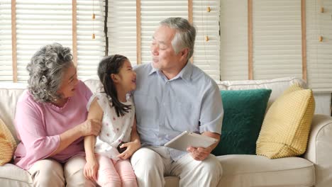 Little-girl-comes-to-hug-her-grandfather-and-grandfather-while-they-playing-tablet.-Kid-playing-with-senior-man-and-woman-with-happy-emotion.-people-with-family,-lifestyle,-technology-concept.