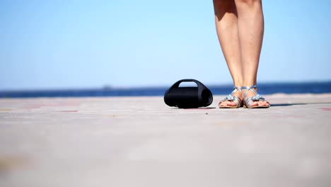 close-up,-mini-music-bluetooth-portable-black-cylinder-wireless-loudspeaker.-female-legs-in-silver-sandals,-with-bright-red-pedicure,-dancing-to-the-music.-on-the-beach,-in-summer