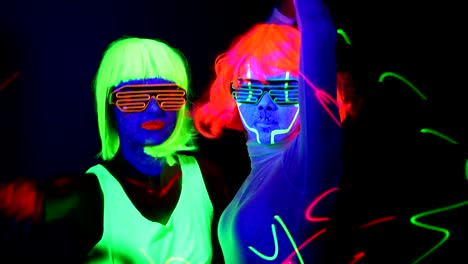 Women-with-laser,-UV-face-paint,-wig,-glowing-glasses,-glowing-clothing-dancing-together-fast-in-front-of-camera,-Half-body-shot.-Caucasian-and-asian-woman.-.