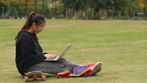 Asian-college-girl-sitting-on-skateboard-using-laptop-in-campus