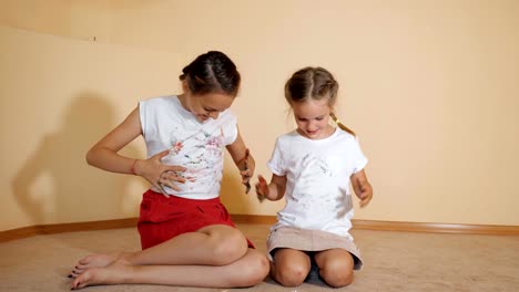 Little-sisters-sitting-on-floor-and-paint-a-t-shirt-with-their-hands