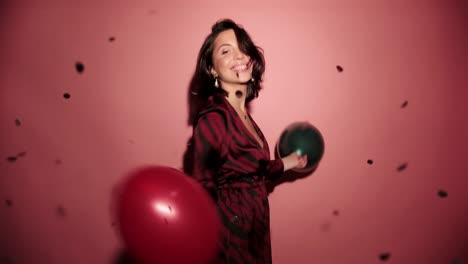 Brunette-happy-woman-dance-with-balloons-and-confetti-in-pink-background-wear-red-dress