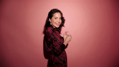 Brunette-happy-woman-give-a-kiss-to-camera-with-confetti-in-pink-background-wear-red-dress