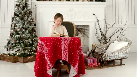 Chico-sonriente-explorando-las-puturas-en-el-libro-sentado-en-la-mesa-cerca-del-árbol-de-Navidad