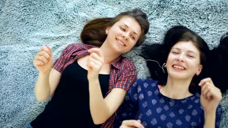 Top-view-of-two-beautiful-girls-in-headphones-listening-music-dancing-and-smiling-while-lying-on-bed