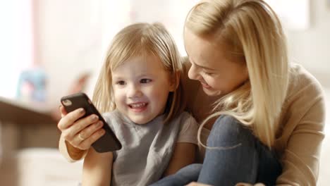 Schöne-junge-Mutter-setzt-sich-mit-Ihrer-kleinen-Tochter-durch-und-zeigt-Ihr-Interessantes-auf-einem-Smartphone.-Das-Kinderzimmer-ist-rosa-und-voll-mit-Spielzeug.