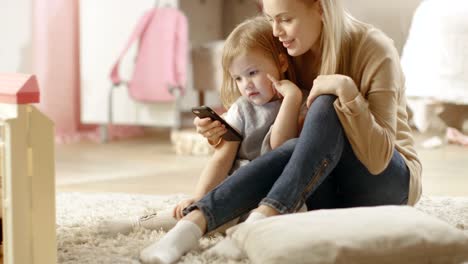 Beautiful-Young-Mother-Sits-with-Her-Little-Daughter-and-Shows-Her-Something-Interesting-on-a-Smartphone.-Children's-Room-is-Pink-and-Full-of-Toys.
