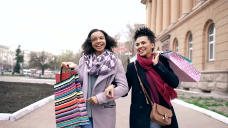 Carro-de-tiro-de-dos-mujeres-de-raza-mixta-atractiva-bailando-y-divertirse-mientras-camina-por-la-calle-con-bolsas-de-la-compra.-Jóvenes-amigos-felizes-caminar-tras-visitar-venta-centro-comercial