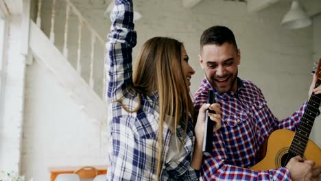 Funny-happy-and-loving-couple-dance-singing-with-tv-controller-and-playing-guitar.-Man-and-woman-have-fun-during-their-holiday-at-home