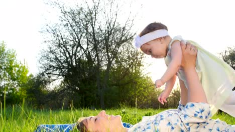 love-is-above-all,-In-the-park,-on-the-grass,-on-the-lawn,-lying-down,-mom,-a-young-blond-woman-playing,-smiling,-with-her-daughter,-a-little-cute-baby,-in-the-sun,-at-sunset.-Im-having-fun,-mom-raising-up-baby,-baby-laughing