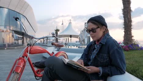 Young-woman-reading-a-book-with-vintage-bike-beside-on-sea-front-at-sunrise