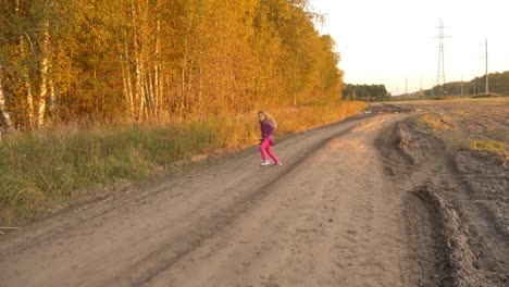Girl-running-and-twisting-on-country-road-autumn-evening.-Outdoors-young-girl-full-length-panning.
