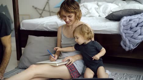 Happy-family-with-cute-adorable-daughter-drawing-in-album-with-pencils-sitting-near-bed-at-home