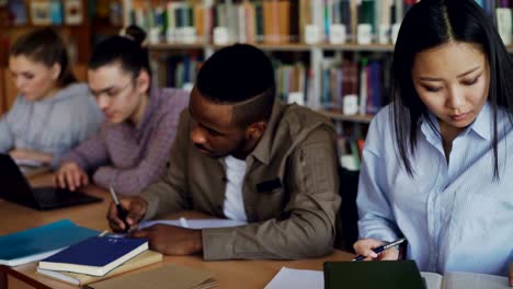 Asiatische-Studentin-auf-Prüfung-vorbereiten-und-Schreiben-von-Notizen-beim-Sitzen-am-Tisch-an-der-Universitätsbibliothek-mit-ihren-Mitschülern-Studium-Bücher-konzentriert