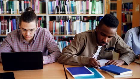 Schwenken-Sie-Schuss-Multi-ethnischen-Gruppe-konzentrierte-Studenten-Vorbereitung-für-die-Prüfung-beim-Sitzen-am-Tisch-in-der-Universitätsbibliothek