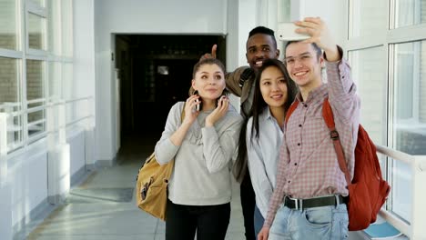 Gruppe-von-multi-ethnischen-Studenten-nehmen-Selfie-auf-Smartphone-Kamera-stehend-im-Flur-der-Universität.-Hipster-Mann-hält-Telefon-und-Freunde-stellen-positiv