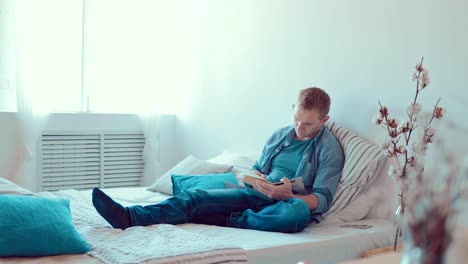 Young-man-reading-book-sitting-on-sofa-at-home