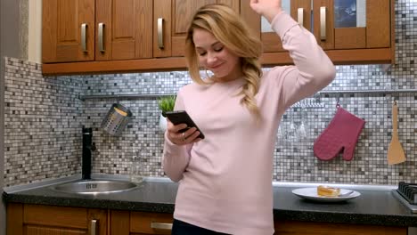 Happy-young-woman-dancing-in-kitchen-listening-to-music-on-smartphone-at-home