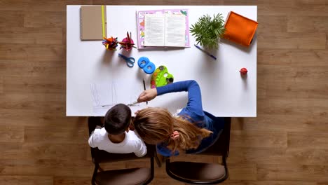 Young-mother-painting-with-son,-sitting-behind-table,-kid-doing-homework-top-shot-parquet-wooden-floor