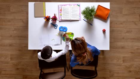 Young-mother-painting-with-son,-explaining-to-kid,-sitting-behind-table,-boy-doing-homework-top-shot-parquet-wooden-floor