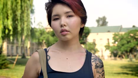 Portrait-of-young-asian-female-student-with-tattoo-standing-with-backpack-and-looking-in-camera-in-park-near-university,-confident