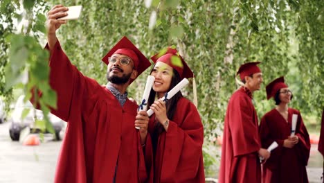 Zwei-mixed-Rennen-Studenten-nehmen-Selfie-mit-Abschluss-Diplom-Mortarboards-und-Kleider-tragen,-Kerl-hält-Smartphone-und-unter-Bild.