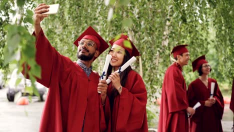 Two-mixed-race-students-are-taking-selfie-with-graduation-diplomas-wearing-mortarboards-and-gowns,-guy-is-holding-smartphone-and-taking-picture.