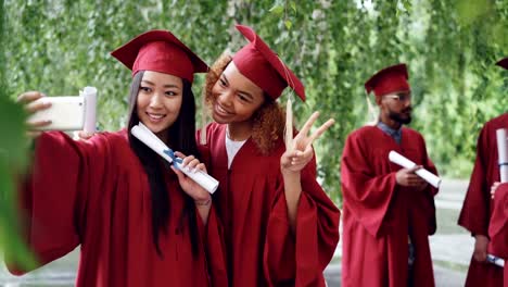 Compañeros-de-dos-chicas-atractivas-están-tomando-selfie-en-graduación-diplomas-de-celebración-de-día,-las-mujeres-jóvenes-están-sonriendo,-posando,-haciendo-gestos-con-las-manos-y-caras-divertidas.