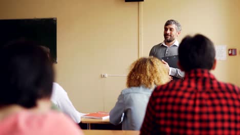 Amable-maestro-está-hablando-con-sus-alumnos-comprobar-conocimientos,-joven-es-levantar-la-mano-y-pregunta,-otros-estudiantes-están-escuchando.-Enseñanza-y-el-aprendizaje-de-concepto.