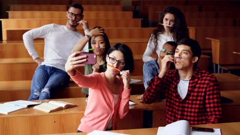 Playful-young-people-are-taking-funny-selfe-using-pens-and-pencils-as-moustache,-posing-and-showing-hand-gestures-thumbs-up-and-v-sign-sitting-in-classroom.