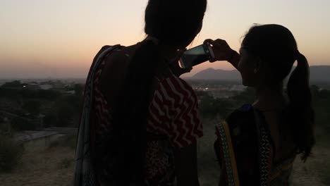 Two-women-silhouette-against-the-setting-sun-mobile-phone-photo-video-teamwork-romantic-viewpoint-love-bonding-together-tourist-rural-setting-friends-handheld-stabilized-lovers-follow--escape-surreal