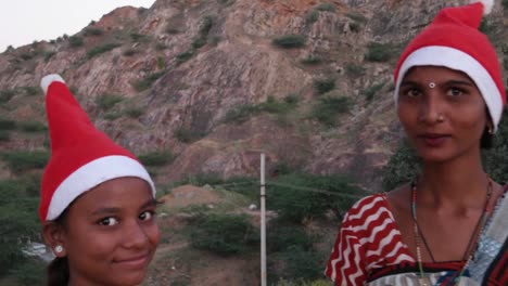 Two-women-in-Santa-hats-and-Indian-traditional-sari-bright-pattern-colorful-dress-summer-tropical-on-a-hill-top-hug-share-point-finger-look-straight-at-camera-sunset-silhouette-laugh-fun-joy-handheld