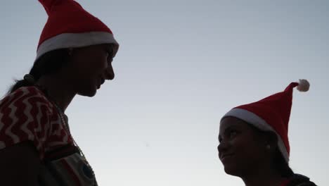 Zwei-Frauen-in-Santa-Hüte-und-traditionellen-indischen-Sari-helles-Muster-buntes-Kleid-Sommer-tropischen-auf-einem-Hügel-oben-Umarmung-Anteil-Punkt-Finger-schauen-Sie-direkt-auf-Kamera-Sonnenuntergang-Silhouette-lachen-Spaß-Freude-handheld