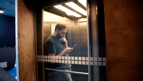 Young-happy-casual-man-riding-transparent-glass-elevator,-door-opens-and-he-walks-out-using-smartphone-shopping-app