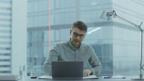 Handsome-Young-Businessman-Sitting-at-His-Desk-Using-Laptop-in-the-Office-Raises-Hands-and-Celebrates-His-Job-Promotion.-In-the-Background-Window-with-Cityscape-View.