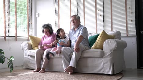 Muchacha-de-los-niños-vienen-a-sentarse-en-el-regazo-de-pareja-senior-y-beso-mejilla-juntos-en-la-sala-de-estar-en-casa.-Concepto-de-excite-de-la-vida,-familia-feliz,-calidad-vida,-salud-mental-y-psicología-positiva.-resolución-de-4-k.