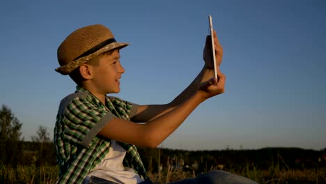 boy-in-a-hat-sits-on-top-and-talks-on-video-communication-using-a-tablet,-outdoors