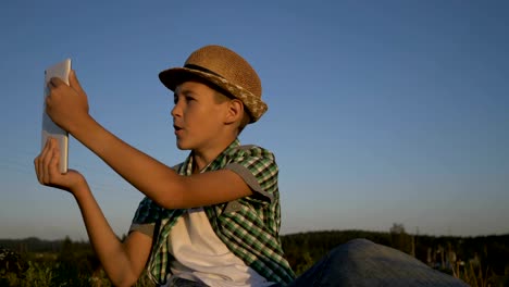 boy-in-a-hat-sits-on-top-and-talks-on-video-communication-using-a-tablet,-outdoors