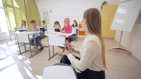 Retrato-de-sonriente-mujer-mujer-de-profesor-durante-la-lección-de-educación-con-alumnos-en-aula-de-escuela-primaria-en-fondo-desenfocado