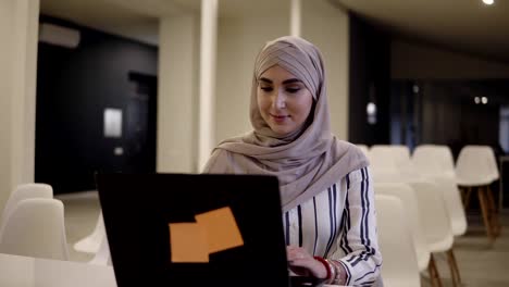 Smiling-confident-muslim-female-wearing-a-hijab-sitting-on-a-white-chair-in-the-modern-conference-hall-online-with-a-laptop-on-the-white-desk