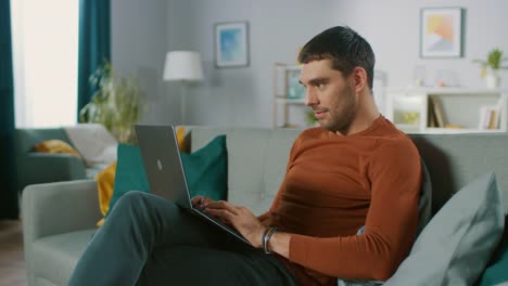 Handsome-Man-Uses-Laptop-Computer-while-Sitting-on-Sofa-at-Home.-Man-Working,-Browsing-Through-Internet-from-His-Cozy-Living-Room.
