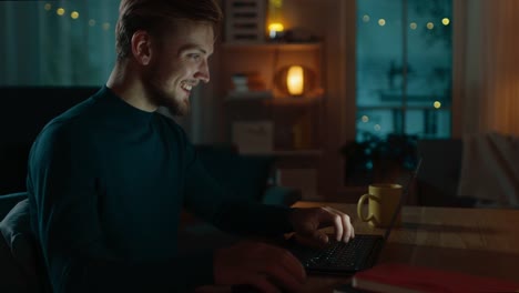 Noche-en-casa:-retrato-de-hombre-guapo-sentado-en-su-escritorio-trabajando-en-un-ordenador-portátil.-Sonriendo-el-Freelancer-trabajando-en-equipo.-Toma-de-vista-de-lado.