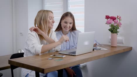 Blonde-and-brunette-women-sitting-at-table-in-kitchen-with-laptop,-watching-funny-videos-online,-fun-indoor