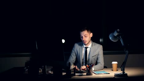 Tired-young-man-in-suit-is-working-on-computer-late-at-night-sitting-in-office-alone-finishing-job-rubbing-his-face-having-headache.-Overwork-and-youth-concept.