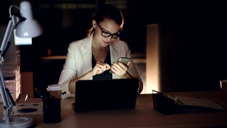 Good-looking-blonde-is-using-smartphone-texting-and-smiling-sitting-in-office-alone-late-at-night.-Modern-gadgets,-hard-working-youth-and-communication-concept.