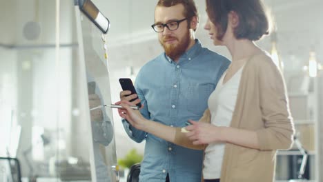 Junger-Mann-und-eine-Frau-diskutieren-Arbeitsprozess-auf-einem-Whiteboard.-Mann-hält-Smartphone.-Frau-stützt-sich-auf-ein-Whiteboard.