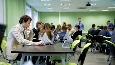 Organizer-of-business-game-sitting-at-the-desk-in-the-classroom-and-using-laptop-during-students-at-the-room-are-doing-practice-and-playing-their-roles-ccording-to-the-rules-of-the-game